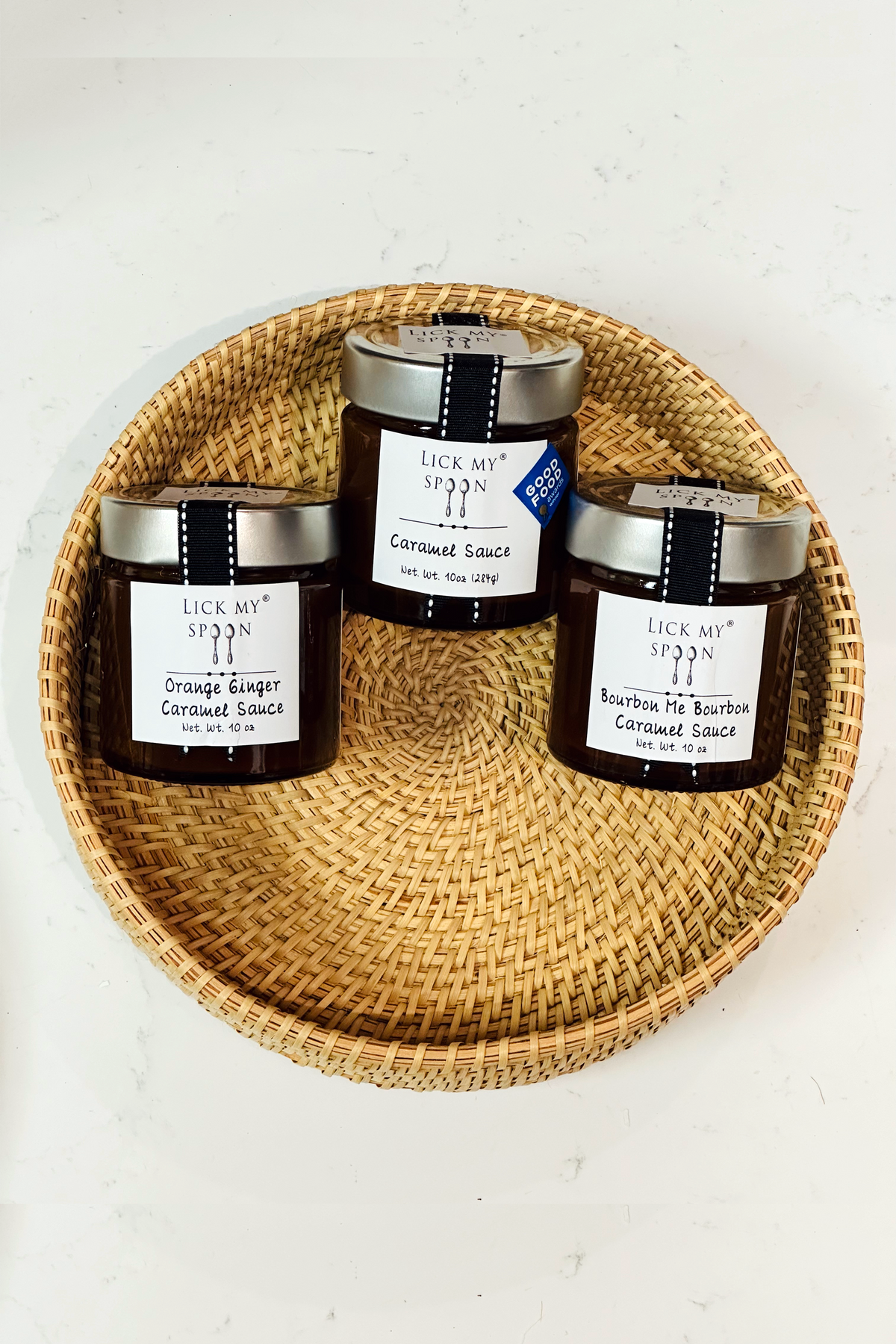 Three jars of "Lick My Spoon" brand caramel sauce displayed side-by-side inside a woven basket 