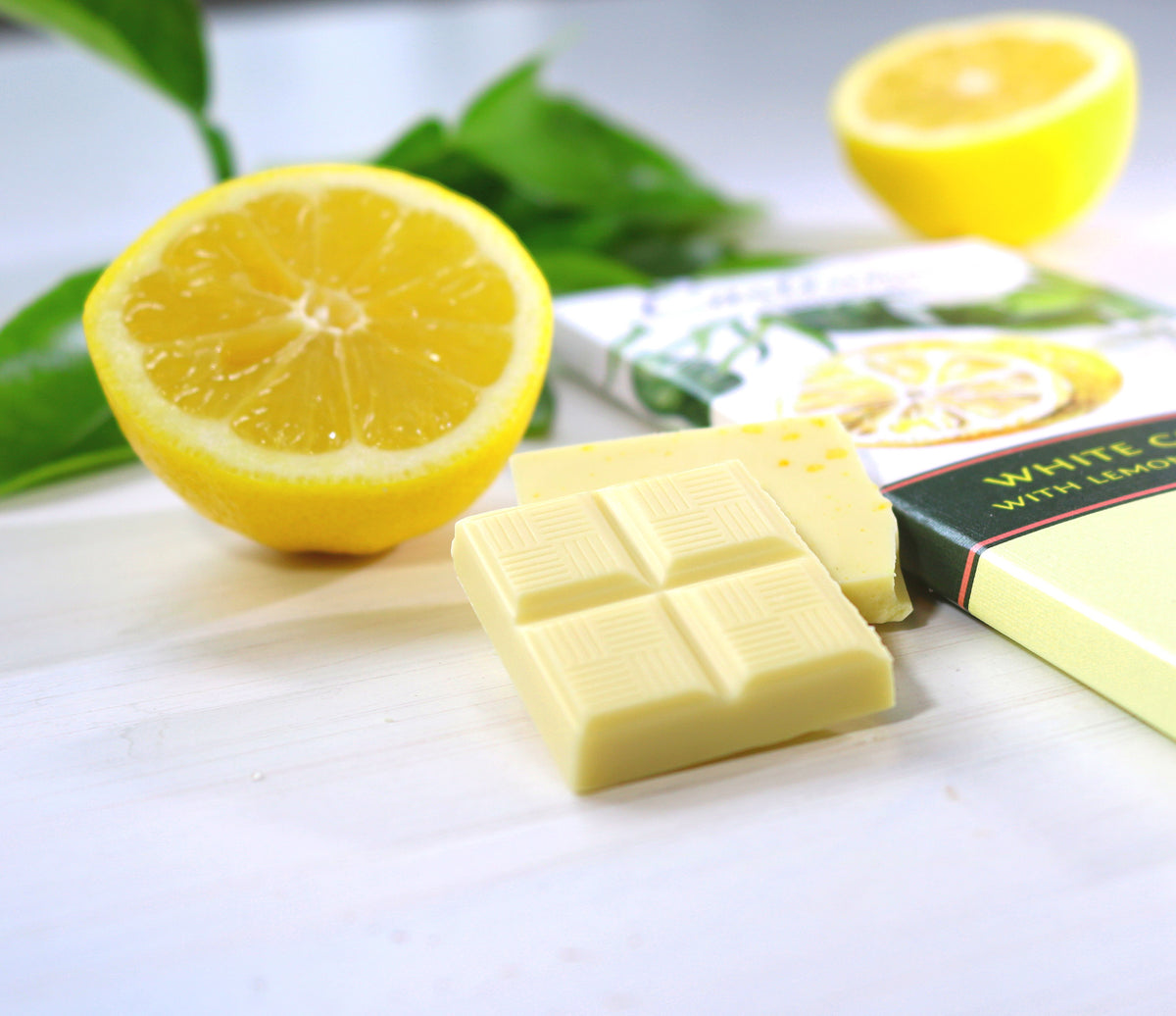 Zoomed in photo of piece of Castronovo brand white chocolate with lemon and lemon sea salt on a white cloth surface, accompanied with sliced lemon 