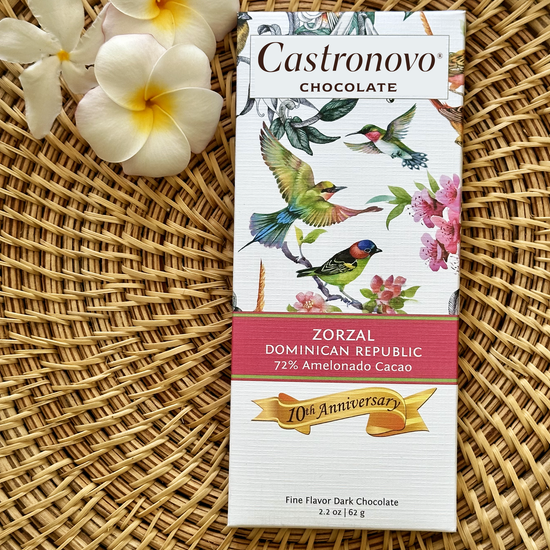 A chocolate bar with a bird and flower design, labeled 'Castronovo Chocolate', resting on a woven mat with a flower in the background.