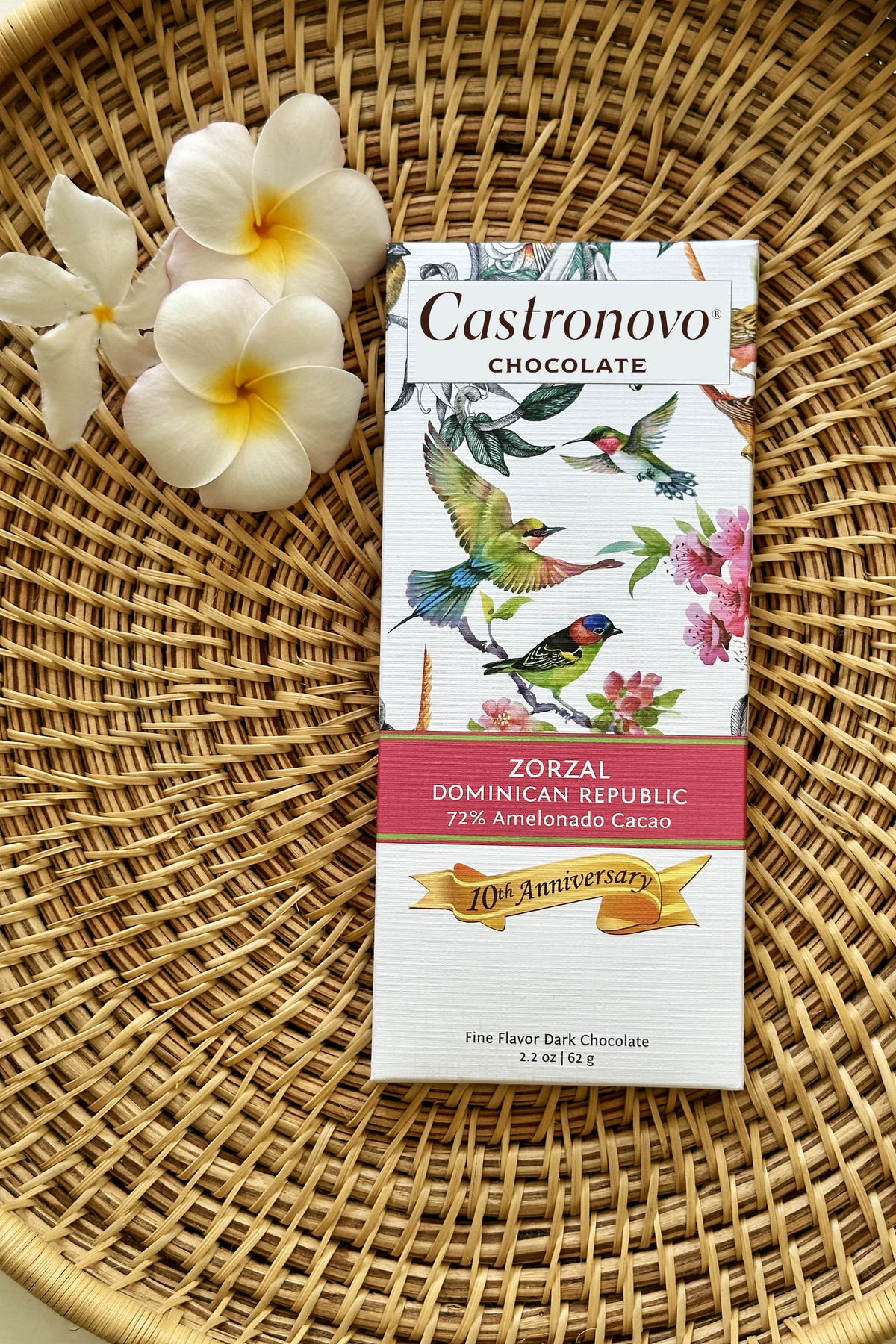 A chocolate bar with a bird and flower design, labeled 'Castronovo Chocolate', resting on a woven mat with a flower in the background.