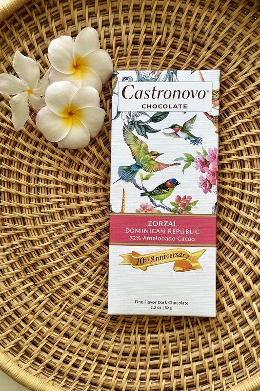 A chocolate bar with a bird and flower design, labeled 'Castronovo Chocolate', resting on a woven mat with a flower in the background.