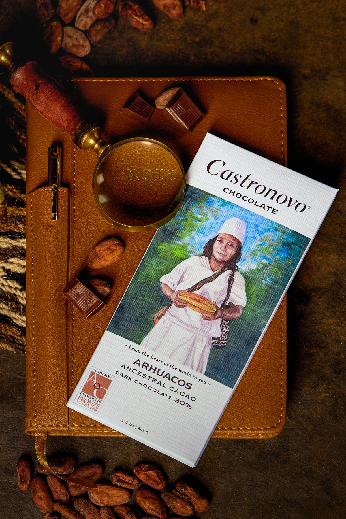 A bar of Arhuacos 80% dark chocolate by Castronovo Chocolate, placed on a surface with cocoa beans and a magnifying glass.